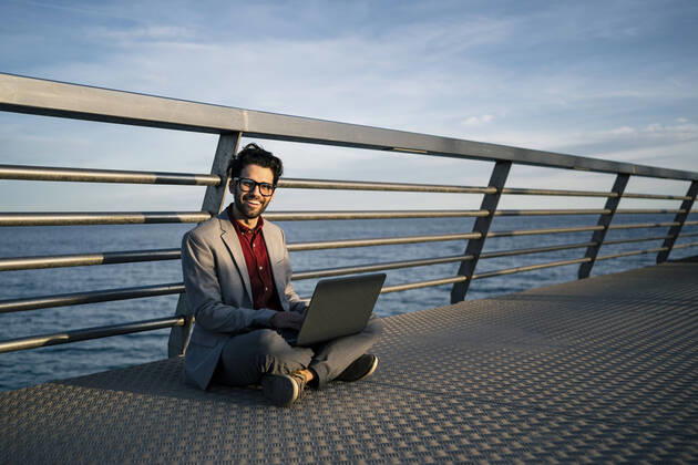 Smiling freelance worker with laptop sitting cross legged on pier model released Symbolfoto RCPF00834