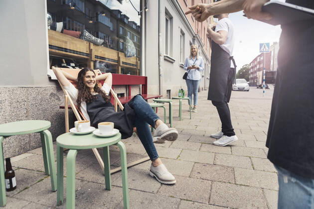 Smiling female owner with hands behind head sitting on deck chair outside coffee shop Sweden, Malmo, model released, property released Copyright: xKentarooxTrymanx , MA85249