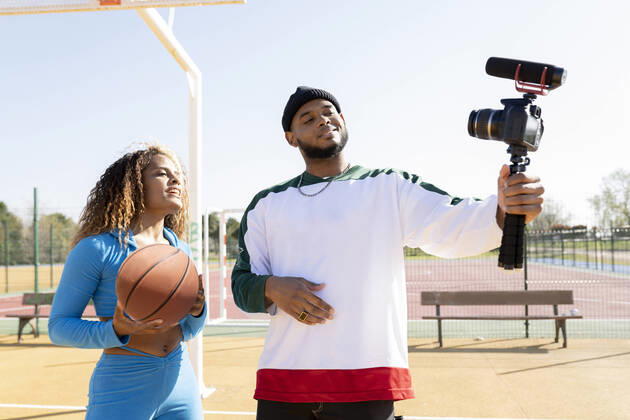 Male holding tripod with camera while vlogging with female friend on sports court model released Symbolfoto JCCMF01722