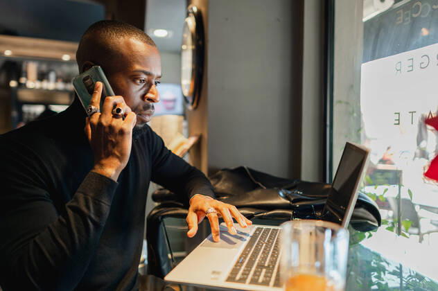 Side view of focused African American male entrepreneur working remotely in cafe while speaking on smartphone and browsing laptop, Model released Copyright: xCarlosxPintaux MR
