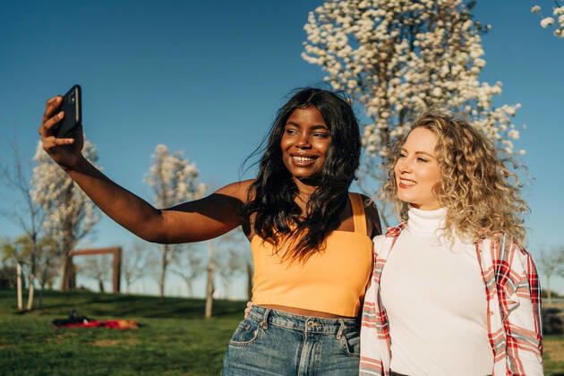 IMAGO Creative: N?chste Generation, Sozial- und klimabewusst Low angle of cheerful multiracial female best friends embracing in spring garden and taking self shot on smartphone on sunny day Copyright: xJakexJakabx Model Released