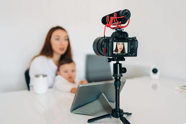 Blurred young mother with cute baby on laps talking and recording video on camera for personal blog while sitting at desk at home Copyright: xJakexJakabx Model Released