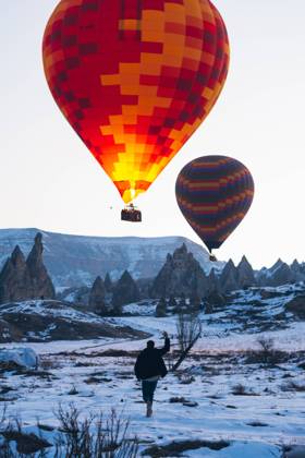 Back view of unrecognizable man in warm clothes with arm raised running to vivid colored air balloon flying up into gray sky over foggy snowy mountain ridge in Turkey, Model released Copyright: xCarlosxForteax MR