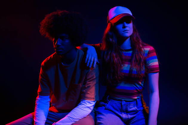 IMAGO Creative: N?chste Generation, Sozial- und klimabewusst Full body of self assured young female in trendy outfit sitting on chair with hand on shoulder of African American boyfriend in dark studio in neon lights, Model released Copyright: xLightsyx MR