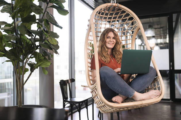 Smiling businesswoman with laptop sitting on swing chair at office cafeteria model released Symbolfoto property released FKF04337