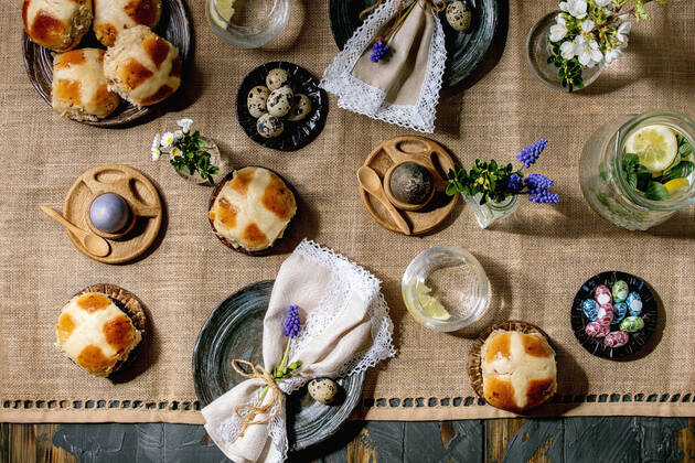 Easter table setting with colored and chocolate eggs, hot cross buns, bouquet flowers, empty ceramic plate with napkin, glass of lemonade drink on wooden table with textile tablecloth. Flat lay NatashaBreen REDA&CO 1028_29_NBR9686