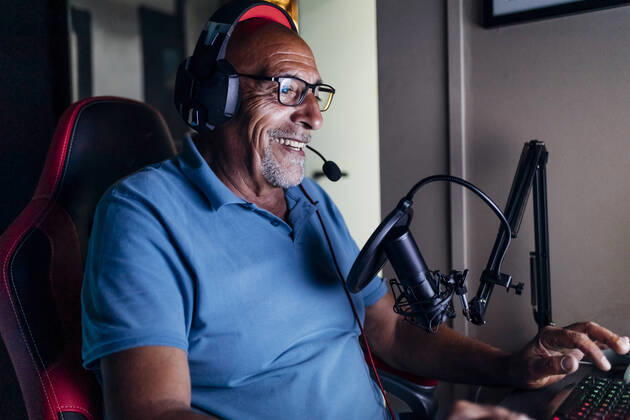 IMAGO Creative, Lifestyle, Gaming Smiling senior man wearing headphones using keyboard while playing video game at home model released Symbolfoto property released MEUF03131
