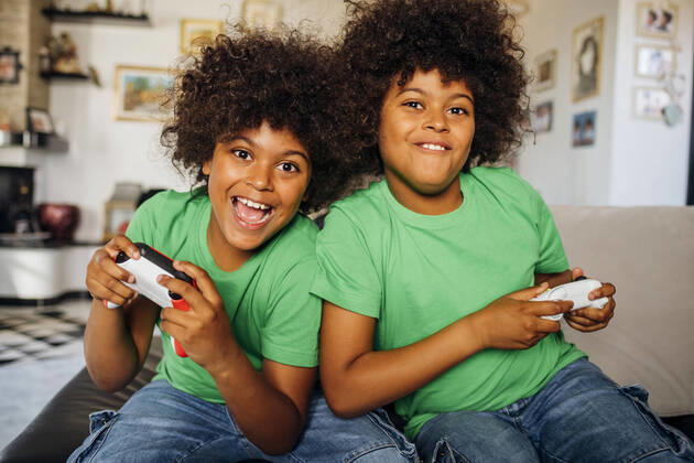 IMAGO Creative, Lifestyle, Gaming Cheerful twin boys with curly hair playing video games sitting on sofa in living room at home model released Symbolfoto property released MEUF03133