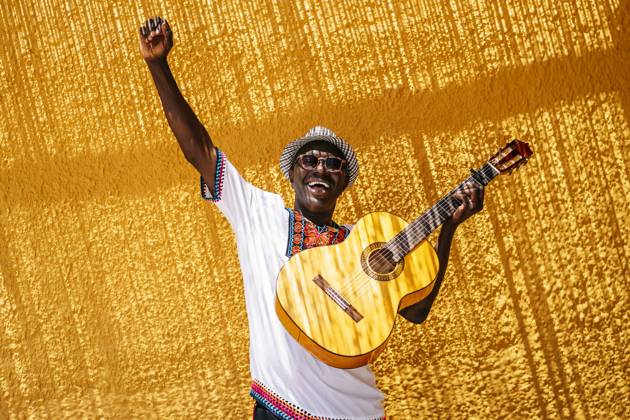 Cheerful man with hand raised carrying guitar in front of yellow wall during sunny day model released Symbolfoto OCMF02148