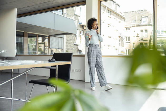 Smiling businesswoman gesturing while talking on mobile phone by window at office model released Symbolfoto property released UUF23265