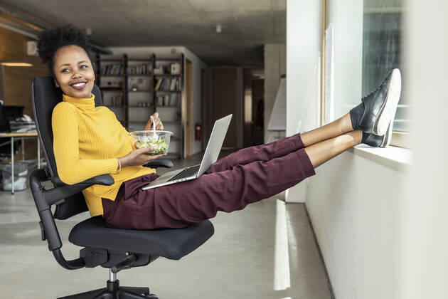 Smiling businesswoman with laptop looking away while having salad on chair at apartment model released Symbolfoto property released UUF23339