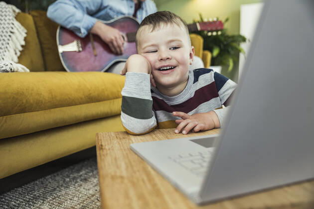 Creative Highlights Symbolbilder Cute boy watching video on laptop while father playing guitar in living room model released Symbolfoto property released UUF23410