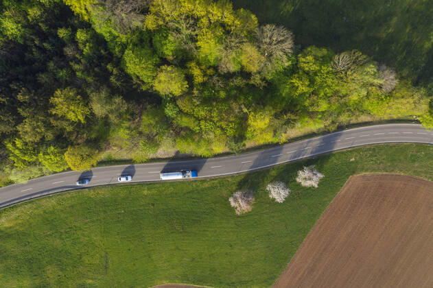 IMAGO Creative: Klimawandel - Zukunft und Innovation Drone view of cars driving along countryside road stretching between green grove and plowed field RUEF03355
