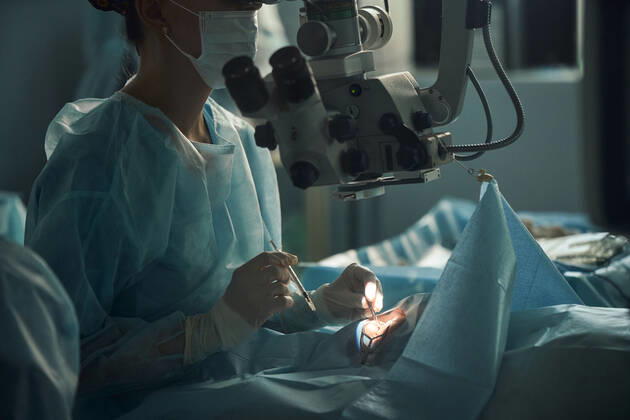 Adult female doctor in sterile mask and ornamental medical cap looking through surgical microscope against crop coworker in hospital, Model released SergeyMironov_Surgery_1.jpg Copyright: xSergeyxMironovx MR