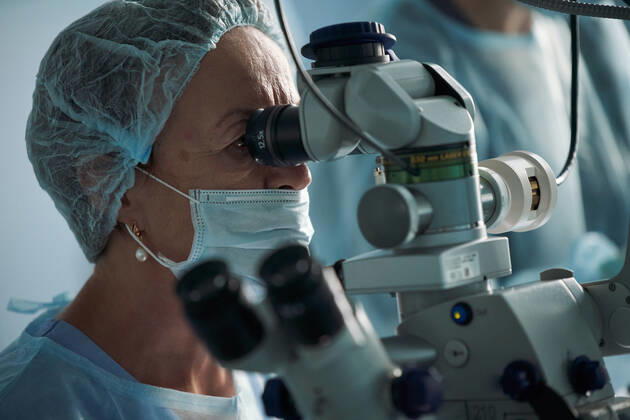 Adult female doctor in sterile mask and ornamental medical cap looking through surgical microscope against crop coworker in hospital, Model released SergeyMironov_Surgery_30.jpg Copyright: xSergeyxMironovx MR
