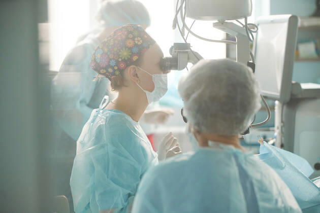 Adult female doctor in sterile mask and ornamental medical cap looking through surgical microscope against crop coworker in hospital, Model released SergeyMironov_Surgery_34.jpg Copyright: xSergeyxMironovx MR