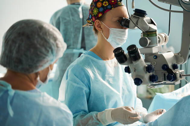 Adult female doctor in sterile mask and ornamental medical cap looking through surgical microscope against crop coworker in hospital, Model released SergeyMironov_Surgery_35.jpg Copyright: xSergeyxMironovx MR