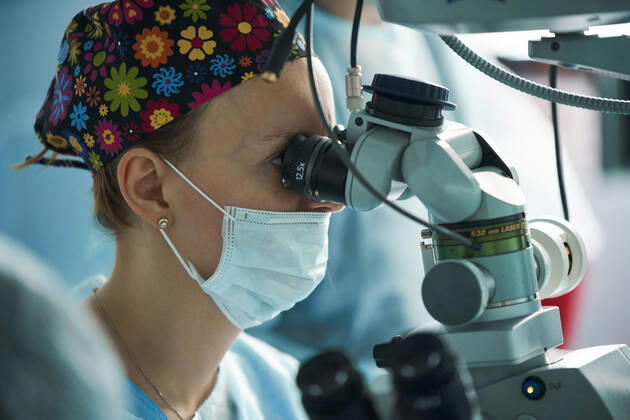 Adult female doctor in sterile mask and ornamental medical cap looking through surgical microscope against crop coworker in hospital, Model released SergeyMironov_Surgery_49.jpg Copyright: xSergeyxMironovx MR