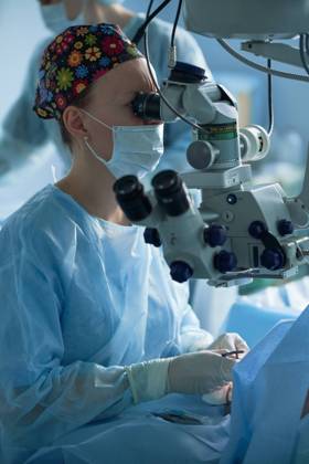 Adult female doctor in sterile mask and ornamental medical cap looking through surgical microscope against crop coworker in hospital, Model released SergeyMironov_Surgery_5.jpg Copyright: xSergeyxMironovx MR