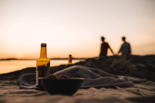 Beer bottle and bowl on picnic blanket while gay couple in background at lakeshore during sunset Sweden, Gothenburg, model released Copyright: xMaskotx , MA88016