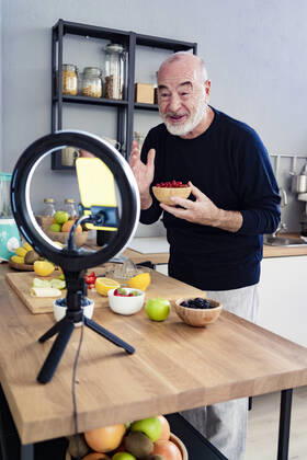 Senior male vlogger with fruit bowl filming through mobile phone at home model released Symbolfoto property released GIOF13790