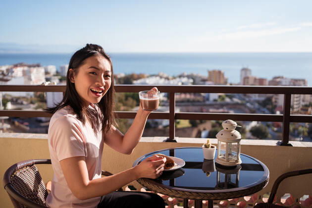 Happy Asian female with glass of hot drink in hand looking at camera while sitting on balcony at table with food during breakfast, Model released JuanAlbertoRuiz_BreakfastAtHome_7.jpg Copyright: xJuanxAlbertoxRuizx MR