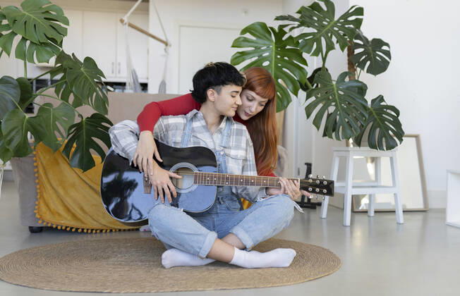 IMAGO Creative: Bildung, Musikerziehung Content lesbian hugging talented female partner playing on acoustic guitar on rug in light living room with green potted plants JoseCarlosIchiro_CoupleWomaenAtLoft_37.jpg Copyright: xJosexCarlosxIchirox Model Released