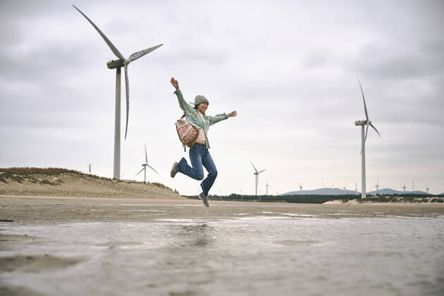 IMAGO Creative: N?chste Generation, Sozial- und klimabewusst Side view of young female in casual clothes with backpack jumping on seaside near windmills on cloudy day LuisGomez_ChristinaOnTheBeach_3.jpg Copyright: xLuisxGomezx Model Released