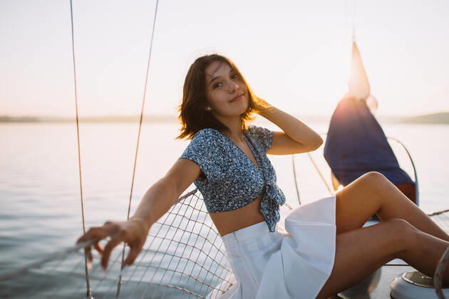 Side view of positive young stylish female traveler with dark hair in trendy crop top and skirt looking at camera while sitting on sailboat during cruise under sunset sky, Model released CamilleGirard_Sailboat_00029.jpg Copyright: xCamillexGirardx MR