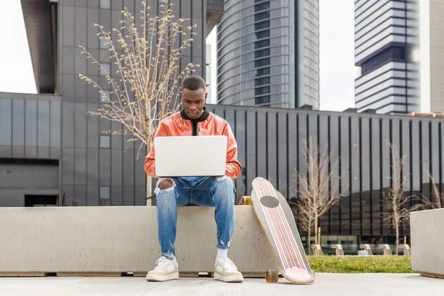 IMAGO Creative: Business - Work-Life Balance Concentrated young African American male freelancer in trendy outfit sitting on stone bench and working remotely on laptop near longboard in city park, Model released IgnacioFerrandiz_ZGenerationStudent_5.jpg Copyright: xIgnacioxFerrandizx MR