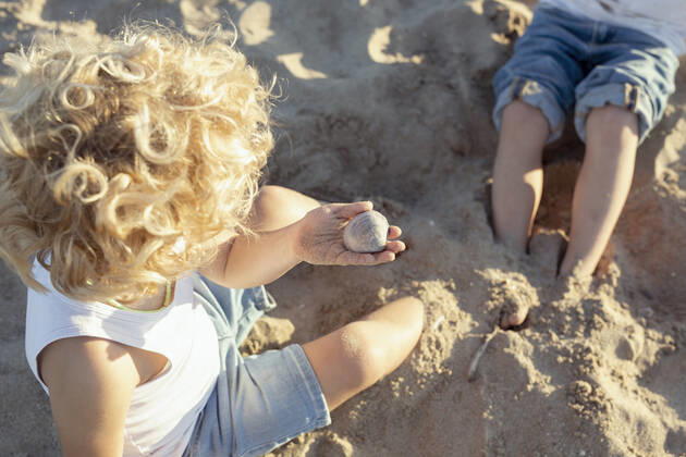 IMAGO Creative, Jahreszeit Sommer, Sommerferien From above of unrecognizable little child with curly blond hair in casual clothes holding pebble while sitting on sandy beach and playing with crop sibling during summer holidays SinuheGorris_EveningWithFriends_00034.jpg Copyright: xSinuhexGorrisx Model Released