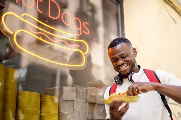 IMAGO Creative, Urban Lifestyle Happy young man eating hot dog leaning on restaurant window model released, Symbolfoto property released, OCMF02415