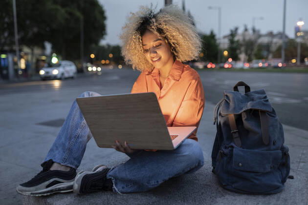 IMAGO Creative: Business, Innovative Technologie-Lösungen Woman using laptop sitting by backpack on sidewalk model released, Symbolfoto, JCCMF06660