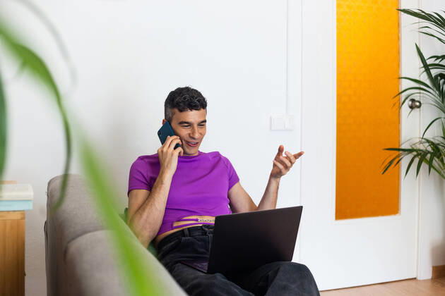 IMAGO Creative: Business, Innovative Technologie-L?sungen Smiling transgender man using phone and laptop sitting on sofa at home, Model released JordiBataller_QueerReality_48.jpg Copyright: xJordixBatallerx MR