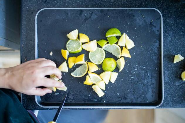  Female hands slicing lemons and limes on a black tray, Top view of female hands carefully slicing lemons and limes on black tray. Woman cutting citric fruit into small pieces forming a heart. Citrus drink preparation, 13.05.2021, Copyright: xKanelbullex Panthermedia30028021.jpg