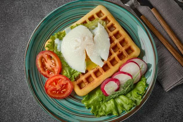  Tasty waffles with poached egg and vegetables on blue plate on grey background. Top view. Table setting, Delicious waffles with poached egg and fresh vegetables on blue glass plate on slate grey background. Top view. Serving food. Table setting, 20.06.2021, Copyright: xDGMx Panthermedia30349019.jpg
