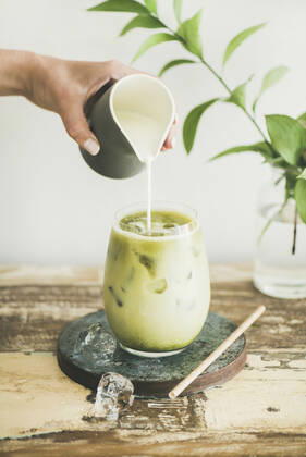 Iced matcha latte drink with milk pouring from pitcher Iced matcha latte drink in glass with coconut milk pouring from pitcher by woman s hand, white wall and plant branches at background. Summer refreshing beverage cold drink Copyright: xAnnaxIvanovax AI130718-4