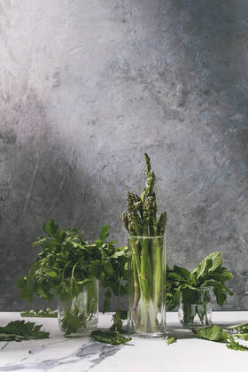 Asparagus ang greens Raw uncooked organic green asparagus, bunch of coriander herbs and fresh mint in glass jars on white kitchen table with grey texture wall as background. Copy space. Copyright: xNatashaxBreenx NB170518-48