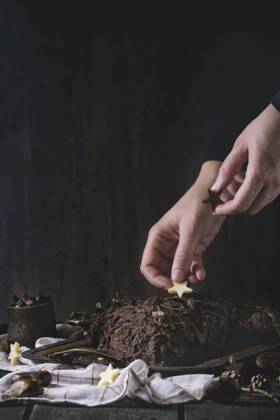 Decorating of christmas chocolate yule log Decorating process of homemade Christmas chocolate yule log by woman s hands with chocolate stars over old wooden table with holly branch and chestnuts. Dark rustic style. With retro filter effect Copyright: xNatashaxBreenx NB271016-3