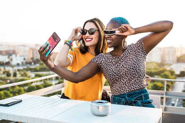 Cheerful multiracial girlfriends in trendy sunglasses smiling sitting at table taking selfie on urban background, Model released RafaCortes_LesbianEthnicCouple_96 Copyright: xRafaxCortesx MR