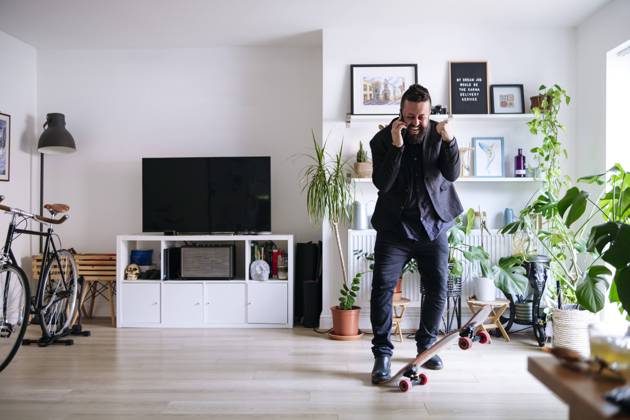 IMAGO Creative: Business, Innovative Technologie-Lösungen Cheerful businessman with skateboard talking on smart phone and clenching fist at home model released, Symbolfoto property released, ASGF02709