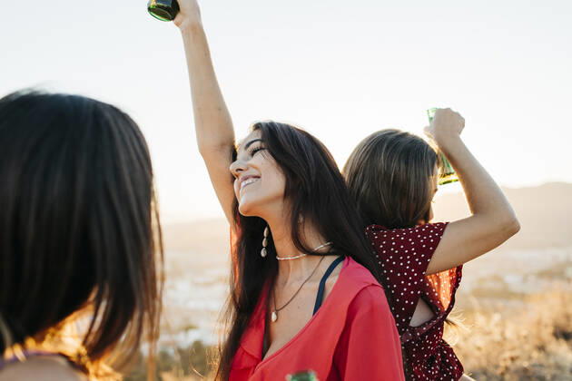 Delighted female friends in stylish outfits with bottles of alcohol beverages standing back to back while dancing on hilltop over town against cloudless sky in sunlight, Model released Lightsy_FriendsMountain_45 Copyright: xLightsyx MR