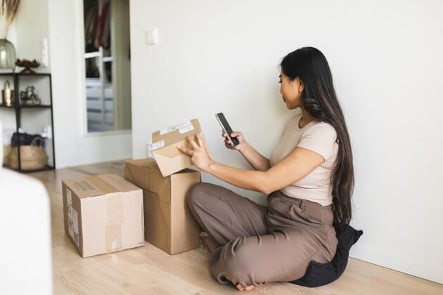 IMAGO Creative: Business, Innovative Technologie-Lösungen Young woman checking and taking picture of delivery package box through smart phone at home model released, Symbolfoto property released, DMMF00232