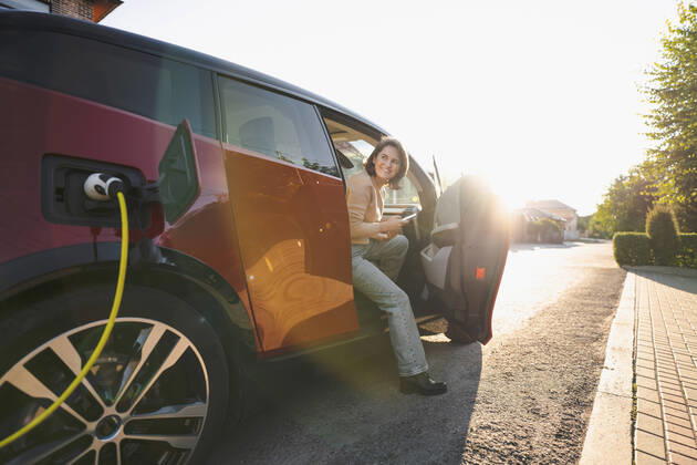 IMAGO Creative: Business, Innovative Technologie-Lösungen Happy woman with mobile phone sitting in electric car at vehicle charging station on sunny day model released, Symbolfoto, EKGF00152