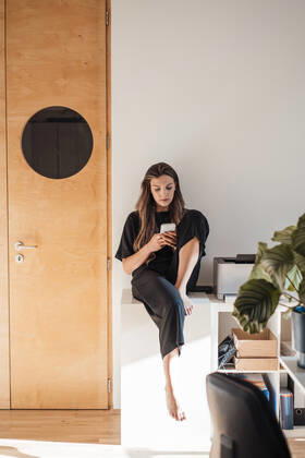 IMAGO Creative: Business, Innovative Technologie-Lösungen Young woman using mobile phone sitting on table at home model released, Symbolfoto property released, JOSEF13433