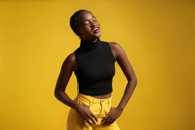 Positive young African American female in casual clothes with braided hair standing near yellow wall with hands in pockets JorgeMierteran_ID10736_325118_053 Copyright: xJorgexMierteranx Model Released