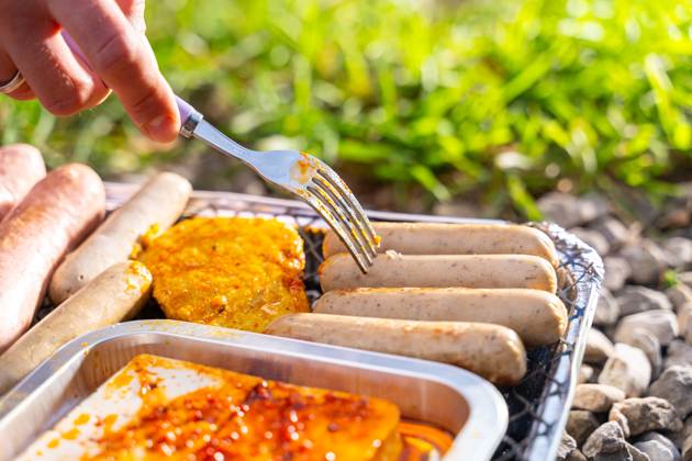 Bavaria, Germany - 2 September 2023: Vegetarian Grilling, Bratwurst Vegan and Vegetarian on a disposable grill. Meat and sausage substitutes. Hand turns grilled food with a fork