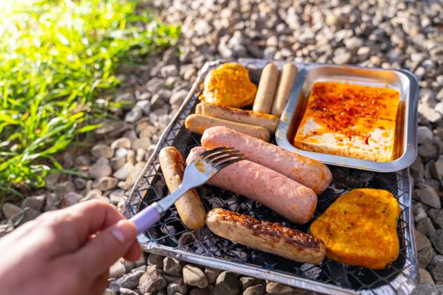 Bavaria, Germany - 2 September 2023: Vegetarian Grilling, Bratwurst Vegan and Vegetarian on a disposable grill. Meat and sausage substitutes. Hand turns grilled food with a fork