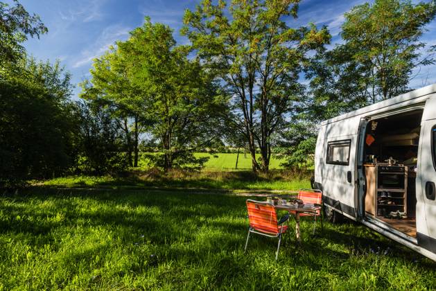 A camper stands between trees on a sunny summers day on a meadow in the countryside, a laid breakfast table with chairs stands in front of the sliding door of the camper Regensburg, Bavaria, Germany In the old town of Regensburg