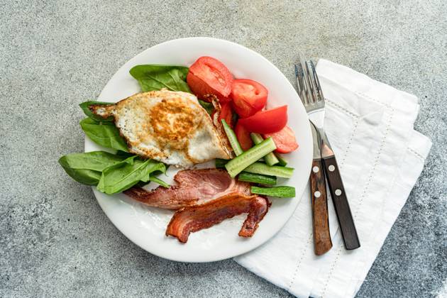 From above of balanced breakfast arrangement on a white plate with fried egg, crispy bacon, fresh spinach leaves, sliced cucumber and wedged tomatoes served with cutlery on grey textured surface Model Released AnnaBogush_ID17558_531810_002 Copyright: xAnnaxBogushx RECORD DATE NOT STATED